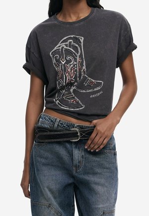 Kvinde iført en falmet sort T-shirt med en detaljeret grafik af cowboystøvler, kombineret med blå denimjeans og et bredt sort bælte med hvide syninger.