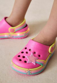 Die Füße eines Kindes in pinken Clogs, verziert mit Regenbogenstreifen und sternförmigen Anhängern, auf einer hellen Oberfläche.