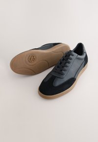 Nizki športni copati z zgornjim delom iz črnega semiša in sivega usnja. Imajo zavezovanje z vezalkami, teksturirano površino in podplat iz gumijastega materiala barve gume.