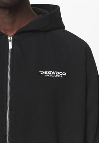 Felpa nera con zip, caratterizzata da una zip argentata, maniche raglan e un logo bianco "PEGADOR ARCTIC CIRCLE" sul petto sinistro.