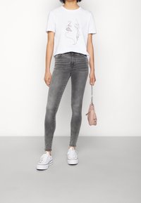 Kvinna i vit t-shirt med abstrakt ansiktsdesign, grå smala jeans, vita sneakers, som håller en liten rosa handväska i kedjehandtaget.