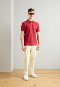 Rotes Poloshirt mit Kragen, hellfarbene Chinos und weiße Sneakers. Modell trägt braune Sonnenbrille. Einfarbiger Hintergrund mit Holzboden.