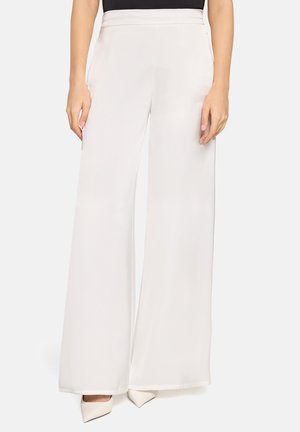 Kobieta w białych, szerokich spodniach z wysokim stanem i bocznymi kieszeniami oraz białych butach na obcasie z ostrym noskiem, stoi na jednolitym tle.