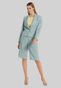 Lichtblauwe linnen blazer met een enkele knoop, gecombineerd met bijpassende knielange shorts en een gele top. Model draagt zilveren sandalen met bandjes.