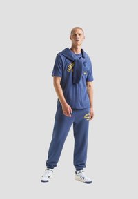 T-shirt in cotone blu da uomo con grafica gialla, abbinato a pantaloni tuta blu e sneakers bianche. Felpa appoggiata sulle spalle.