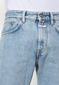 Vaqueros de mezclilla azul claro con cintura alta, diseño clásico de cinco bolsillos y cierre de botón metálico, con un ligero desgaste y textura sutil.