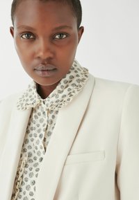 Weißer Blazer über einer hellen, gemusterten Bluse mit Rüschensaum. Das Model hat kurze Haare, einen neutralen Hintergrund und einen konzentrierten Ausdruck.