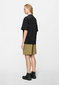 Άτομο που στέκεται πλάγια φορώντας χαλαρό μαύρο πουκάμισο με κοντά μανίκια, χακί σορτς και μαύρες μπότες αστραγάλου σε λευκό φόντο.