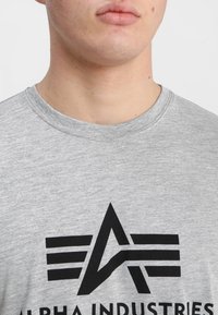 Grå crew-neck t-shirt lavet af blødt stof med et fremtrædende sort grafisk logo med vinklede linjer og tekst på brystområdet.