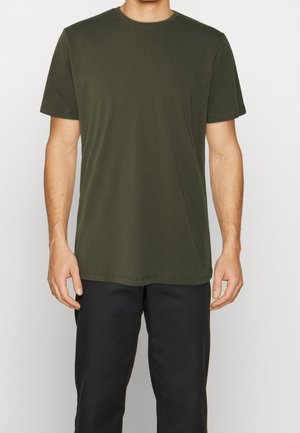 Man draagt een effen olijfgroen T-shirt met korte mouwen en zwarte broek, staat tegen een effen lichte achtergrond.