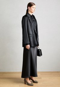 Čierny oversized blazer s textúrovaným povrchom, skombinovaný s dlhou čiernou sukňou. Olivovo zelené podpätky a malá čierna kabelka dotvárajú celkový vzhľad.