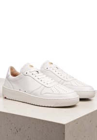 Bílé kožené tenisky s hladkým povrchem, perforovanou špičkou, plochými tkaničkami a texturovanou podrážkou. Zlatý detail loga na jazyku.