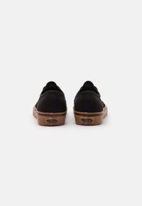 Scarpe slip-on nere in tela con suole in gomma, caratterizzate da una zeppa marrone chiaro e dettaglio del logo sul tallone. Texture liscia e design minimalista.