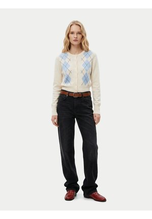 Femme portant un cardigan blanc avec un motif losanges bleu, un jean noir avec une ceinture marron et des chaussures en cuir rouges, debout devant un fond uni.