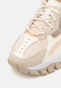 Beige and cream athletic shoe with mesh and suede upper, textured accents, white speckled midsole, and a rugged brown treaded outsole.