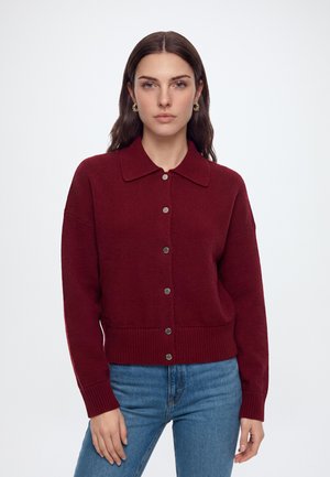 Femme portant un pull en maille bordeaux boutonné avec col et un jean bleu, debout devant un fond blanc uni.