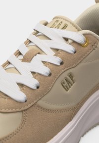 Μπεζ και καφέ αθλητικό παπούτσι με λευκά κορδόνια, χρυσή τρύπα για κορδόνια και λογότυπο "GAP" χαραγμένο στο πλάι και στη γλώσσα, με ραμμένες λεπτομέρειες.