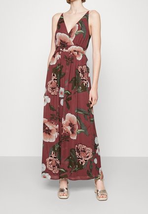 Femme portant une robe longue sans manches bordeaux avec un grand imprimé floral rose et des sandales dorées à talons, debout sur un fond uni.