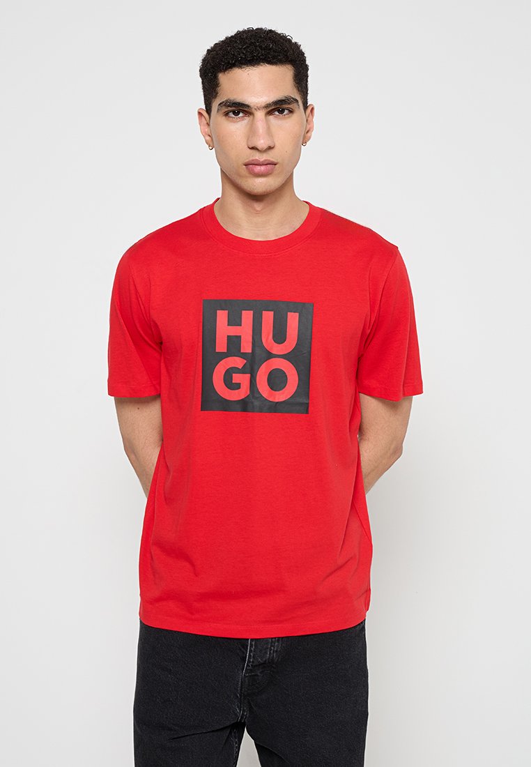 Camiseta de algodón roja con un gráfico cuadrado negro que presenta el texto "HU GO" en letras negritas. Diseño de manga corta y cuello redondo.