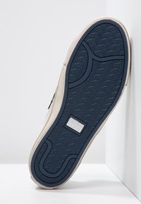 Baskets beige avec une semelle en caoutchouc texturée de couleur bleu marine. Elles présentent un design épuré, une tige lisse et un patch logo près de la semelle.