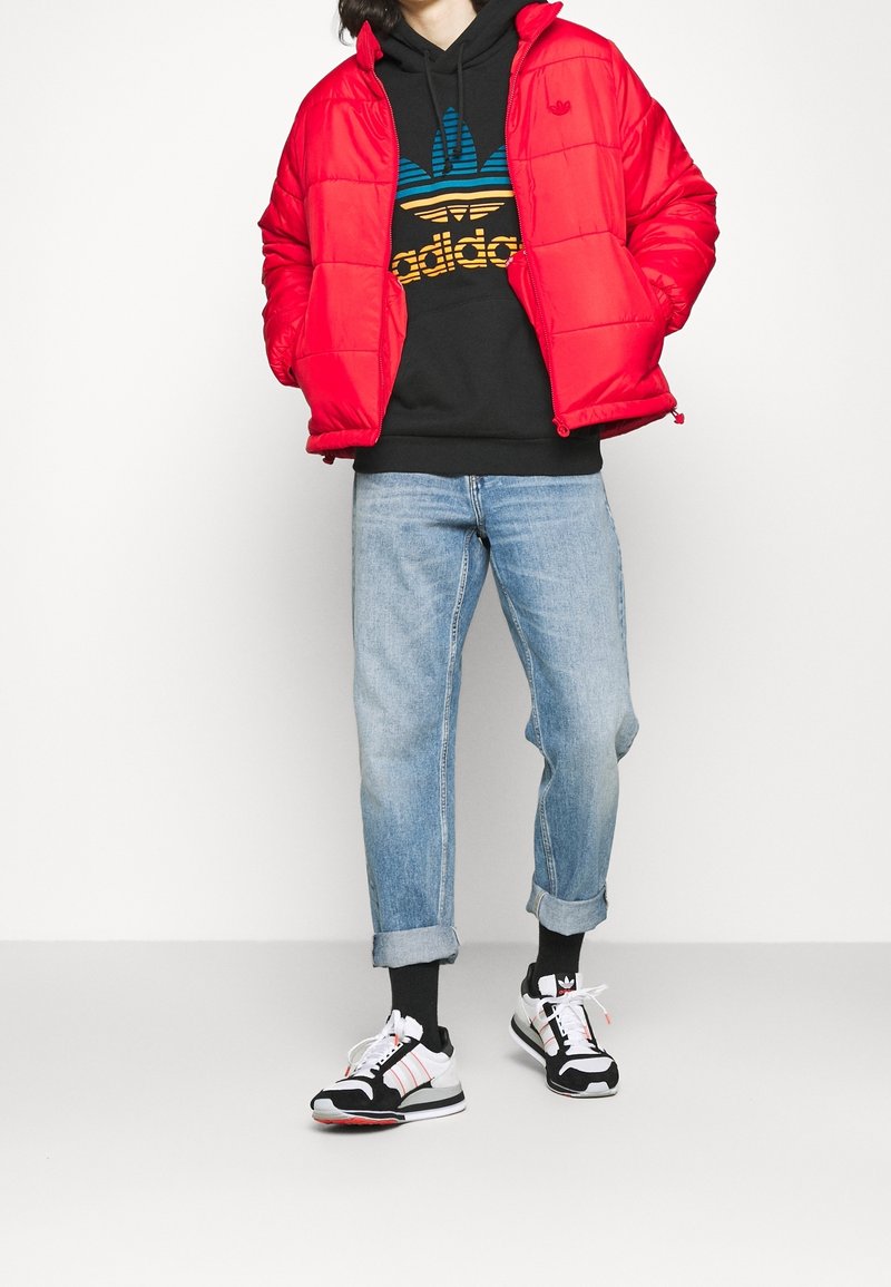 Červená péřová bunda přes černou mikinu s oranžovým a modrým logem Adidas, světle modré rolled-up džíny a černobílé tenisky.