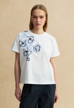 Joven mujer con una camisa blanca de manga corta con bordado floral azul y negro en el hombro izquierdo, combinada con pantalones negros.