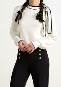 Pull blanc avec accents noirs, manches longues et deux nœuds en ruban noir. Pantalon noir taille haute avec détails de boutons dorés.