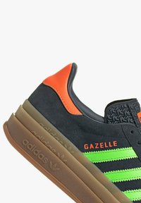 Sneaker nera in suede con strisce verdi e accenti arancioni. Presenta un tallone testurizzato, suola in gomma e il marchio 'GAZELLE'.