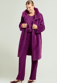 Abrigo de piel sintética púrpura con un cuello ancho, llevado sobre un conjunto púrpura a juego. Presenta un ajuste holgado y una tela texturizada.