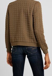 Bruine bomberjack met een gestructureerd quiltpatroon, geribbelde boorden en zoom, gedragen over donkerblauwe spijkerbroek. Heeft een ontspannen pasvorm en een casual ontwerp.