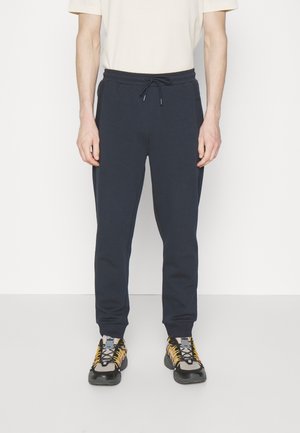 Man draagt marineblauwe joggerbroek met elastische tailleband en trekkoord, gecombineerd met beige en zwarte sneakers met gele veters.