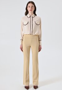 Beige Bluse mit Knopfleiste und zwei Brusttaschen, in Burgunderrot besäumt; beiges, tailliertes Hosenpaar; braune spitze Schuhe mit Schleifen.