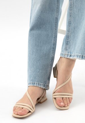 Voeten met beige sandalen met lage hak en gekruiste bandjes, gecombineerd met lichtblauwe spijkerbroek op een witte achtergrond.