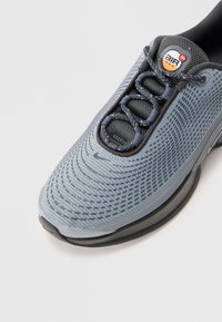 Chaussure de sport grise avec un dessus en mesh texturé, un bout arrondi et des accents noirs. Inclut une unité de rembourrage visible dans la semelle.