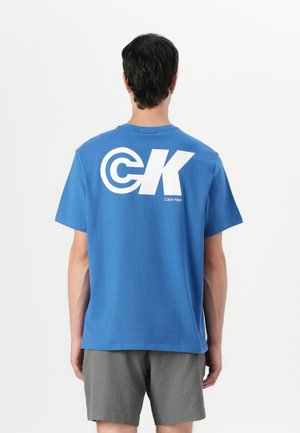 Blaues Baumwoll-T-Shirt mit kurzen Ärmeln, das ein großes weißes "CK"-Logo und den Text "Calvin Klein" auf der Rückseite zeigt. Texturierte graue Shorts sind darunter sichtbar.