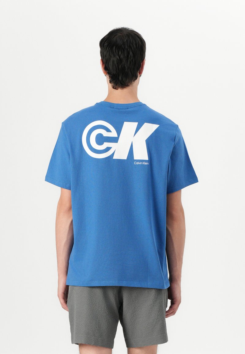 Mėlynas medvilninis marškinėliai trumpomis rankovėmis, su dideliu baltu "CK" logotipu ir "Calvin Klein" tekstu ant nugaros. Apačioje matomos tekstūruotos pilkos šortai.