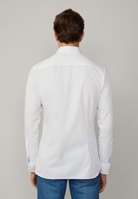 Chemise blanche à manches longues avec une coupe ajustée, col à boutons et accents bleus sur les poignets. Texture lisse, fabriquée en tissu léger.