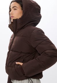 Brązowa kurtka puchowa z wysokim kołnierzem i kapturem, z pikowaną strukturą, bocznymi kieszeniami oraz elastycznymi mankietami zapewniającymi dopasowanie.
