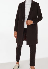Manteau en mélange de laine marron avec des revers crantés, boutons doubles et poches latérales, associé à un pantalon en velours côtelé marron foncé et des mocassins clairs.