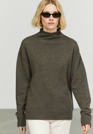 Femme blonde portant des lunettes de soleil ovales noires, un pull à col roulé ample gris foncé et un pantalon clair sur un fond uni.