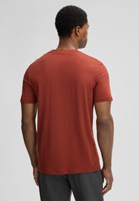 Man staat met zijn rug naar de camera, gekleed in een roestkleurig T-shirt met korte mouwen en donkergrijze broek, tegen een eenduidige achtergrond.