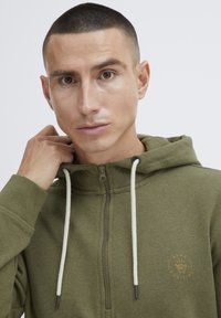 Felpa verde con zip e coulisse bianche, presenta un piccolo logo dorato "Stay Positive" nella zona sinistra del petto. Tessuto morbido.