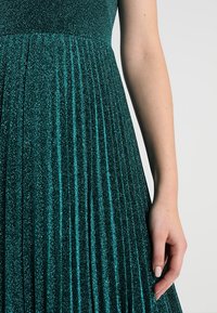 Bras d'une femme à côté d'une robe plissée pailletée bleu sarcelle avec une taille cintrée sur un fond clair uni.