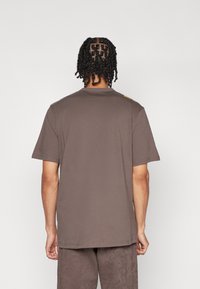 T-shirt en coton brun à manches courtes et col rond. Présente un petit logo jaune sur l'épaule. Texture lisse, coupe décontractée.