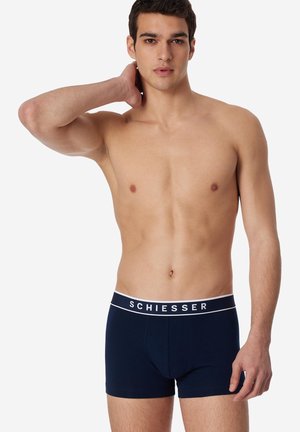 Oberkörperfreier junger Mann, der dunkelblaue Schiesser-Boxershorts trägt, mit der linken Hand den Nacken berührt und vor einem einfachen weißen Hintergrund steht.
