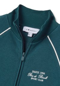 Chaqueta de cremallera en color aguamarina con un cuello acanalado, detalles de bordes blancos y texto bordado "PARIS 1996 Rue de Rivoli SPORT CLUB" en el frente.