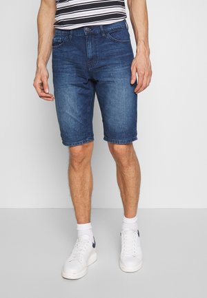Mann trägt blaue Denim-Shorts mittlerer Länge, weiße Socken, weiße Sneakers und ein schwarz-weiß gestreiftes Hemd vor einem einfachen grauen Hintergrund.