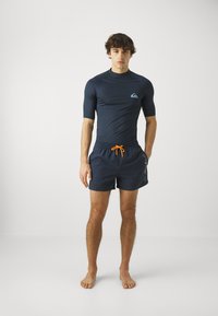 Sininen rash guard t-paidat, jossa on lyhyet hihat ja logo rinnassa, yhdistettynä tummansinisiin uimashortseihin, joissa on oranssi nauha ja sivuominaisuuksia.