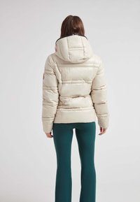 Giacca a vento beige chiaro con cappuccio, caratterizzata da sezioni trapuntate orizzontali e una toppa di gomma sul lato sinistro della spalla. Pantaloni verdi.