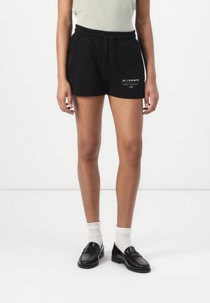 Person som har på seg svarte shorts med snor og hvit logo på venstre lår, hvite sokker og svarte loafers, står mot hvit bakgrunn.